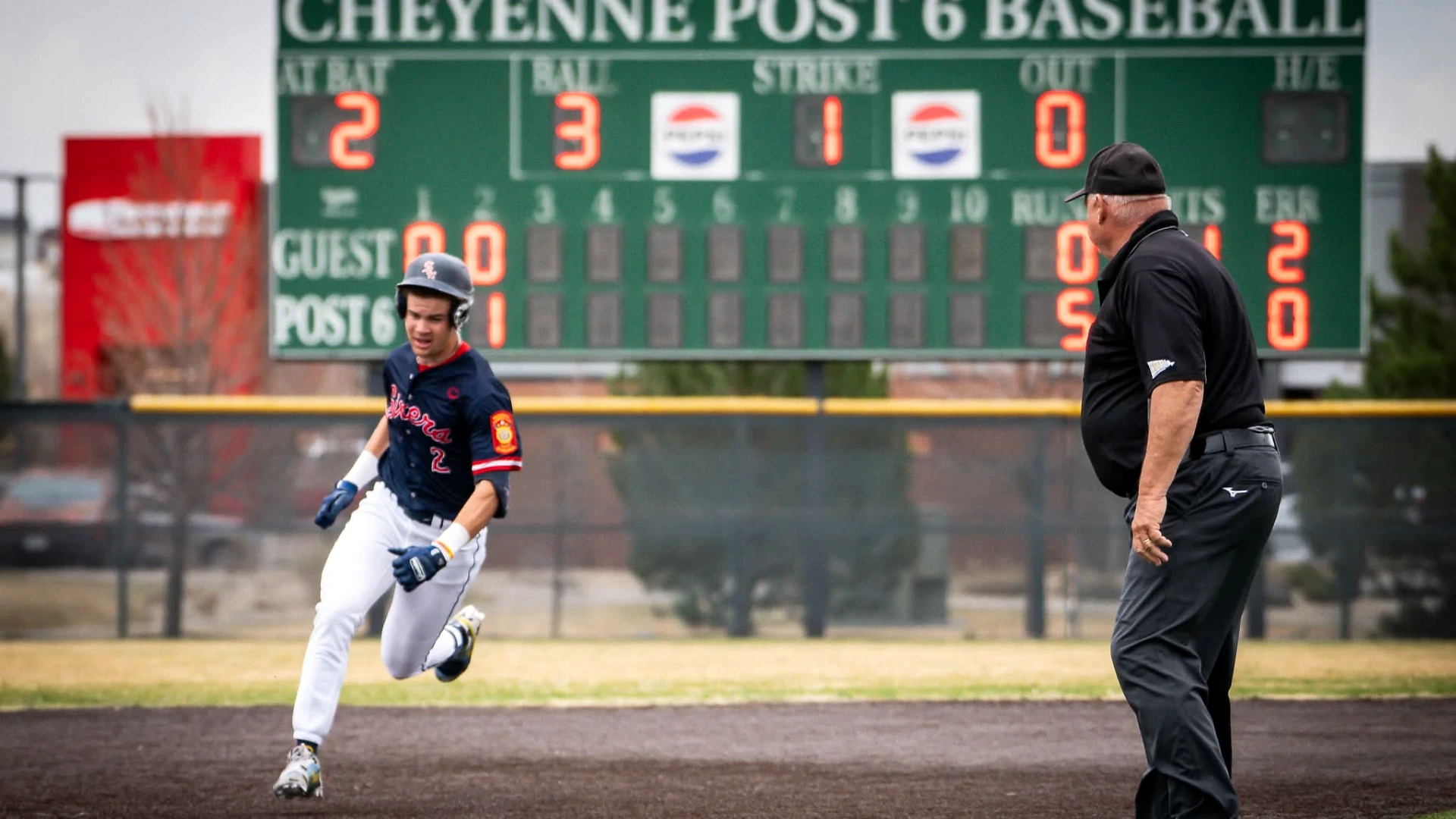 A baseball player sprints past an umpire on the field in front of a Cheyenne Post 6 Baseball scoreboard, showcasing the on-field results of dedicated 1-on-1 sports performance training at Lumen Athletics in Cheyenne, WY.