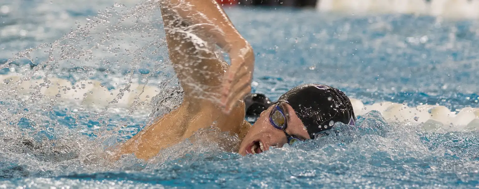 A competitive swimmer in a black cap and goggles powers through blue pool water, one arm lifting above the surface amid energetic splashes.