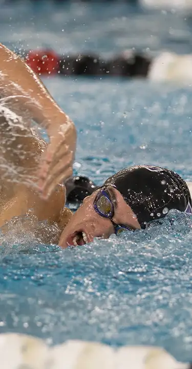 A competitive swimmer in a black cap and goggles powers through blue pool water, one arm lifting above the surface amid energetic splashes.