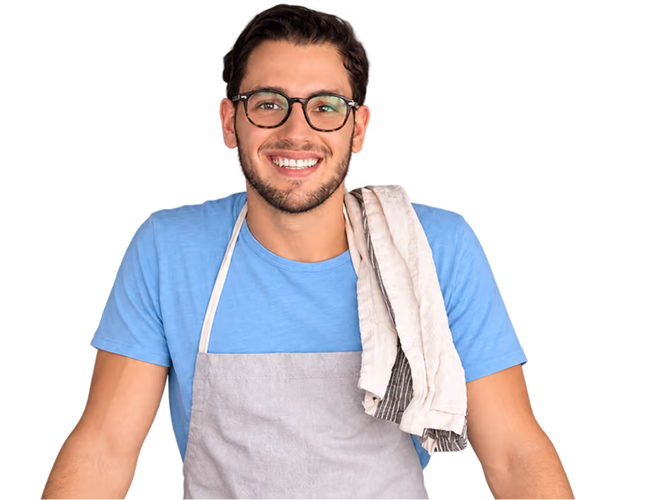 Hombre joven sonriente con gafas, camiseta azul y delantal gris, con un paño blanco y gris sobre el hombro.