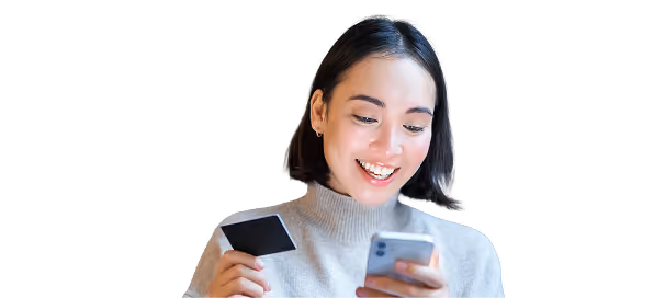 Mujer joven con suéter gris sonriendo, sosteniendo una tarjeta de crédito negra en una mano y un teléfono móvil en la otra, mientras mira la pantalla.