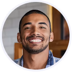 Hombre joven sonriendo con barba de candado y camiseta azul de cuadros, fondo interior desenfocado.
