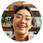 Mujer sonriente con lentes transparentes y cabello oscuro, en un fondo interior con plantas y estantes.