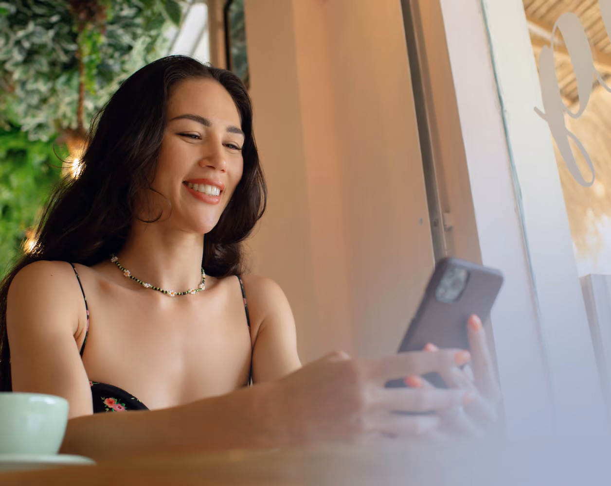 Mujer sonriendo mientras mira un teléfono móvil al aire libre con un edificio alto detrás y luz solar brillante.