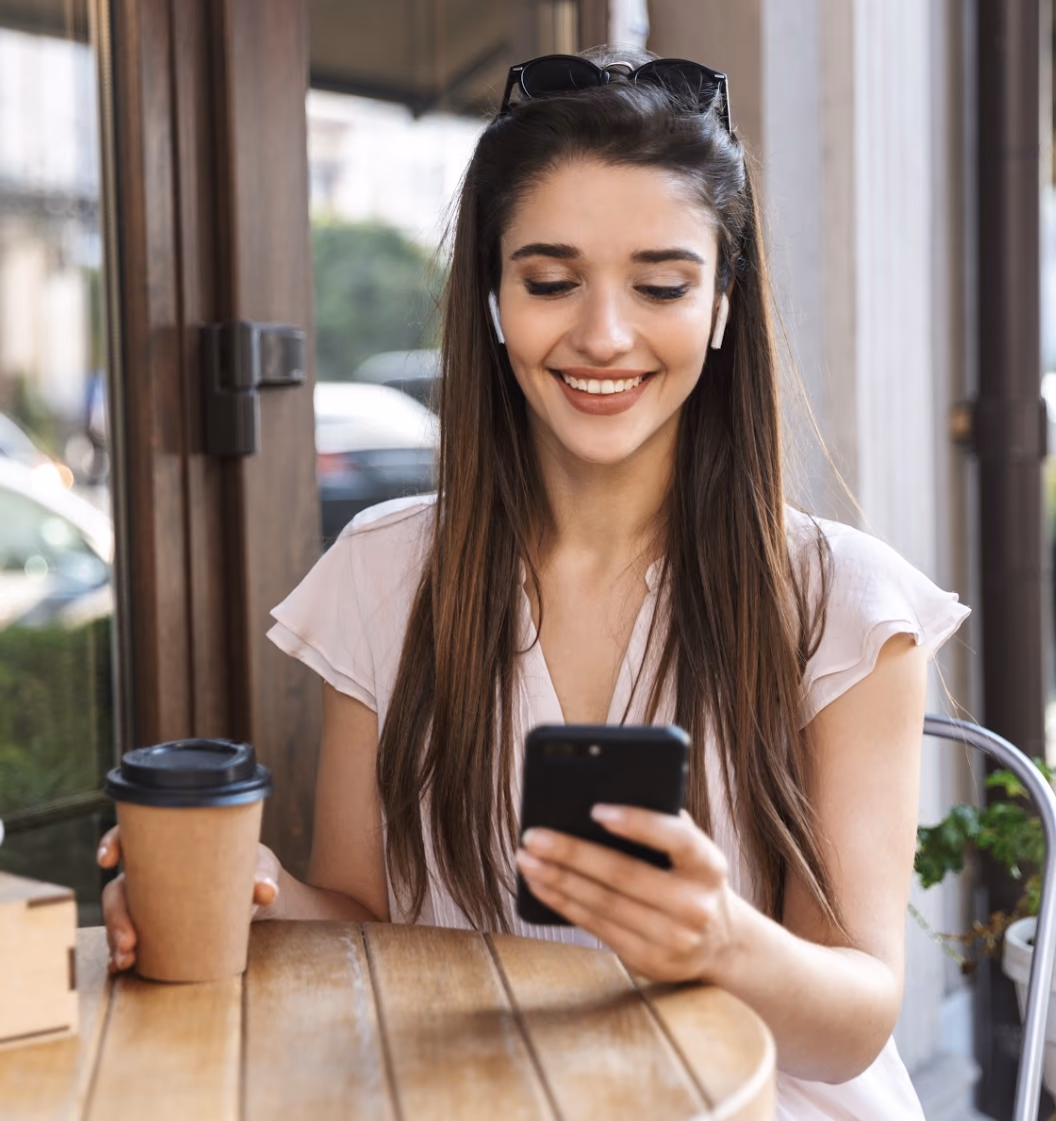 Mujer sonriendo mientras usa su celular en la mesa de un café, con un vaso de café en la mano.