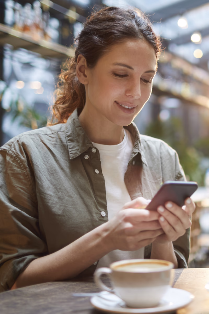 Mujer sonriendo mientras usa su celular en una cafetería, con una taza de café sobre la mesa.