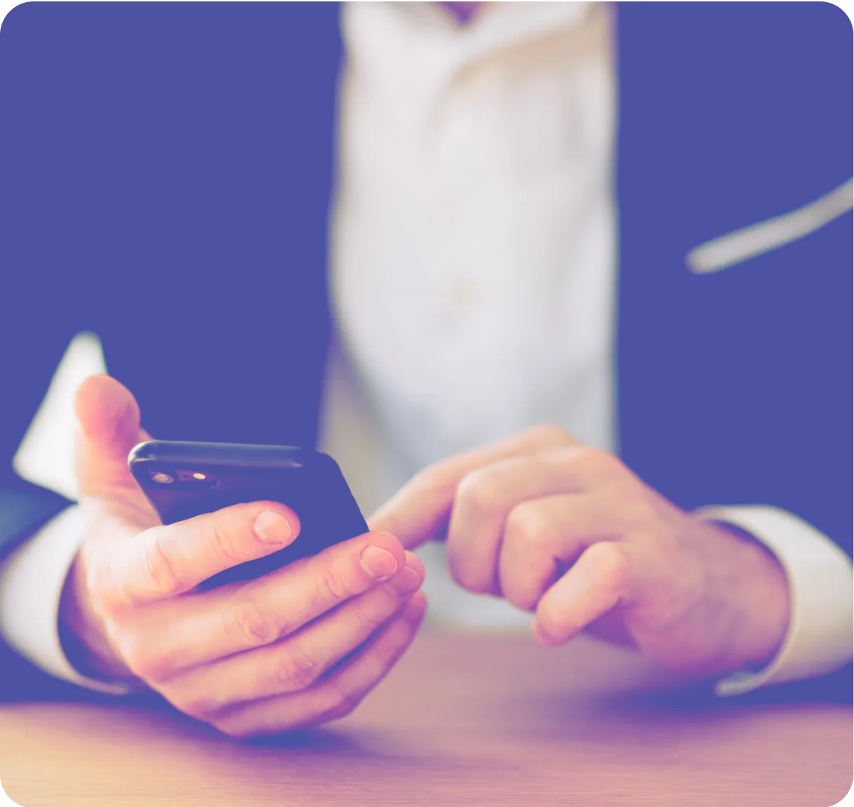 Person in a suit using a smartphone, holding it in one hand while tapping the screen with the other hand over a wooden surface.