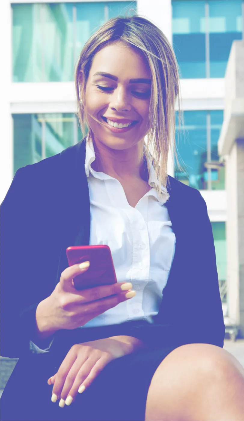 Smiling woman in business attire sitting outdoors and looking at her red smartphone, with a modern glass building in the background.