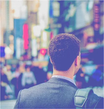 Man in a gray suit with a shoulder bag viewed from behind, standing in a busy city street with blurred people and colorful signs.
