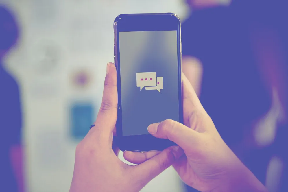 Two hands holding a smartphone displaying a messaging app icon with two speech bubbles and ellipsis indicating typing.