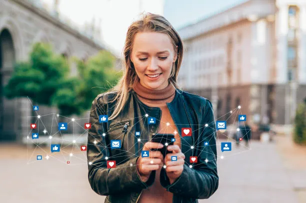 Young woman standing outdoors in an urban area, smiling while using a smartphone with floating social media icons like thumbs-up, hearts, and messages around her.