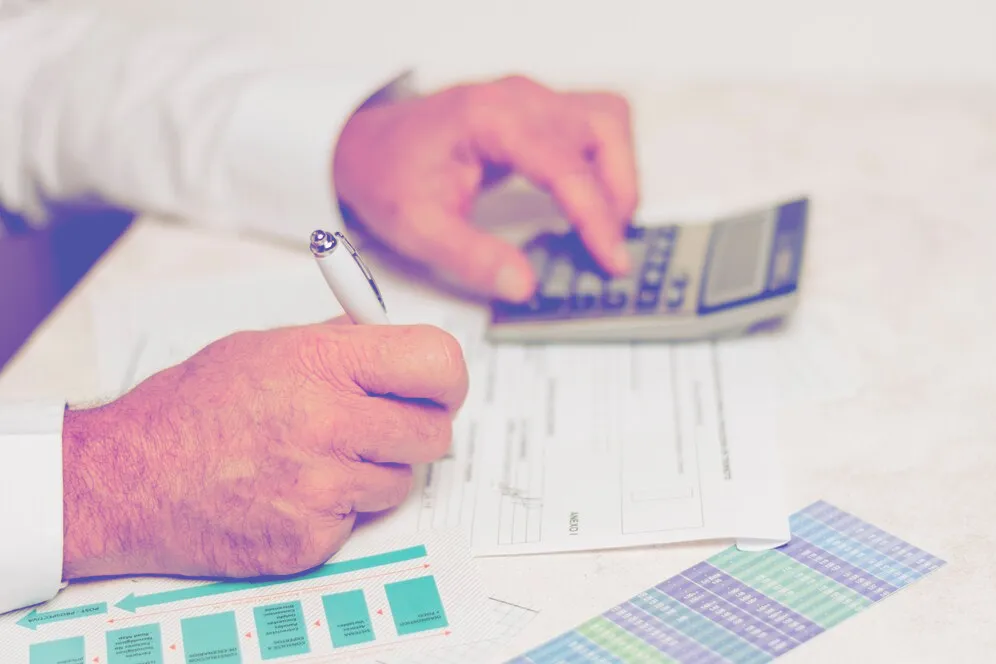 A person using a calculator with one hand and writing on a document with a pen in the other hand, with charts and graphs on the table.