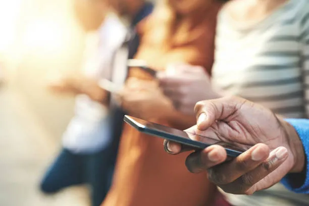 Close-up of diverse people standing in a row using smartphones, with hands and phones in focus and faces blurred.