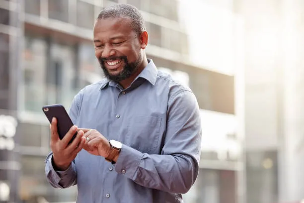 Smiling man wearing a light blue button-up shirt looking at his smartphone outdoors in an urban setting with modern buildings in the background.