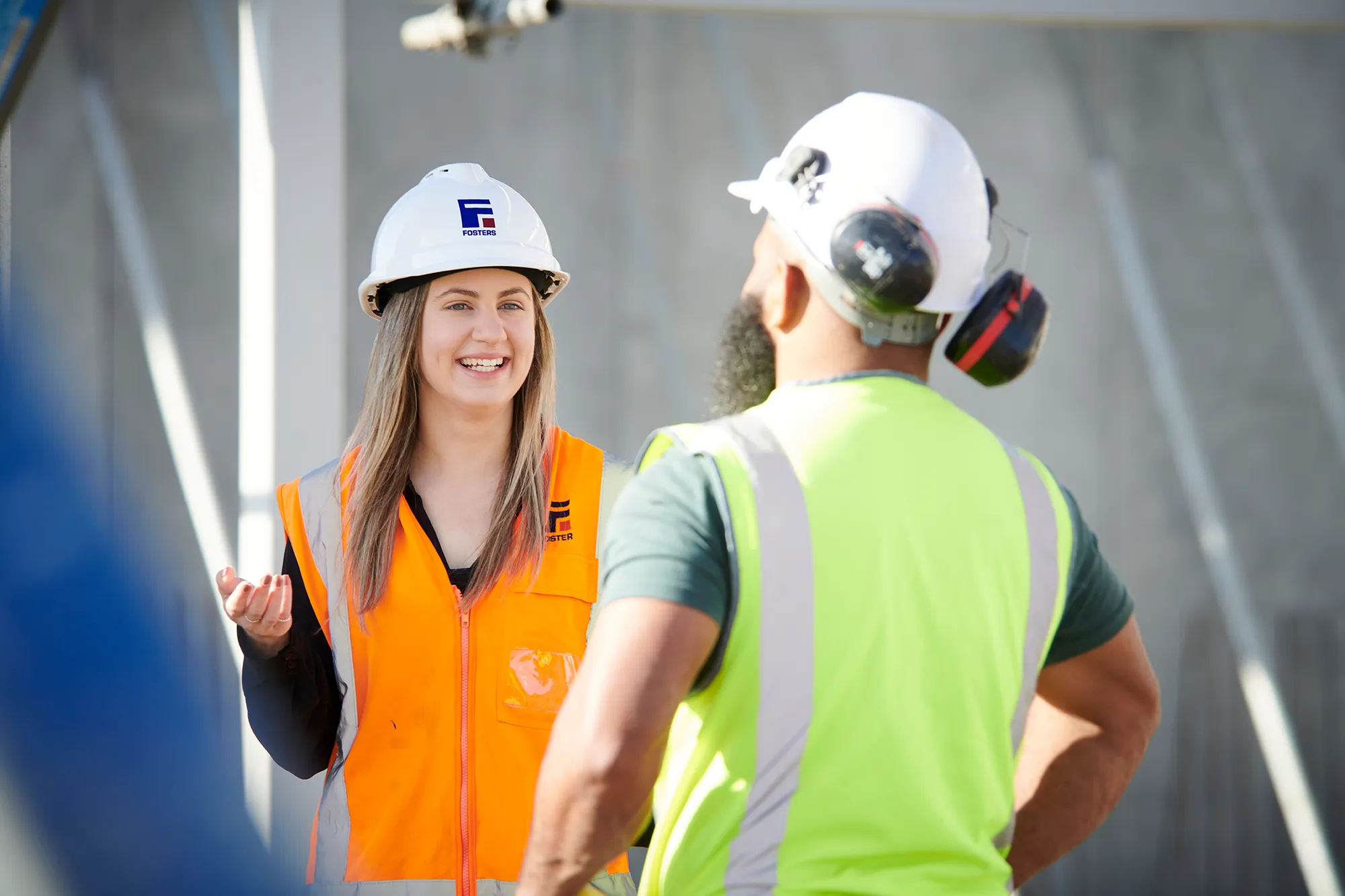 Two construction workers wearing hard hats and safety vests talking on site.