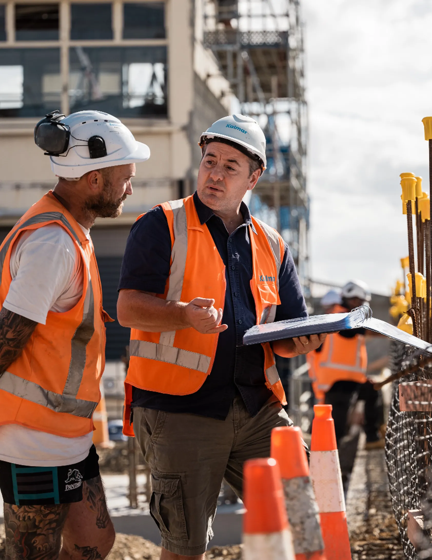 Two construction workers wearing white helmets and orange safety vests discussing plans at a site.