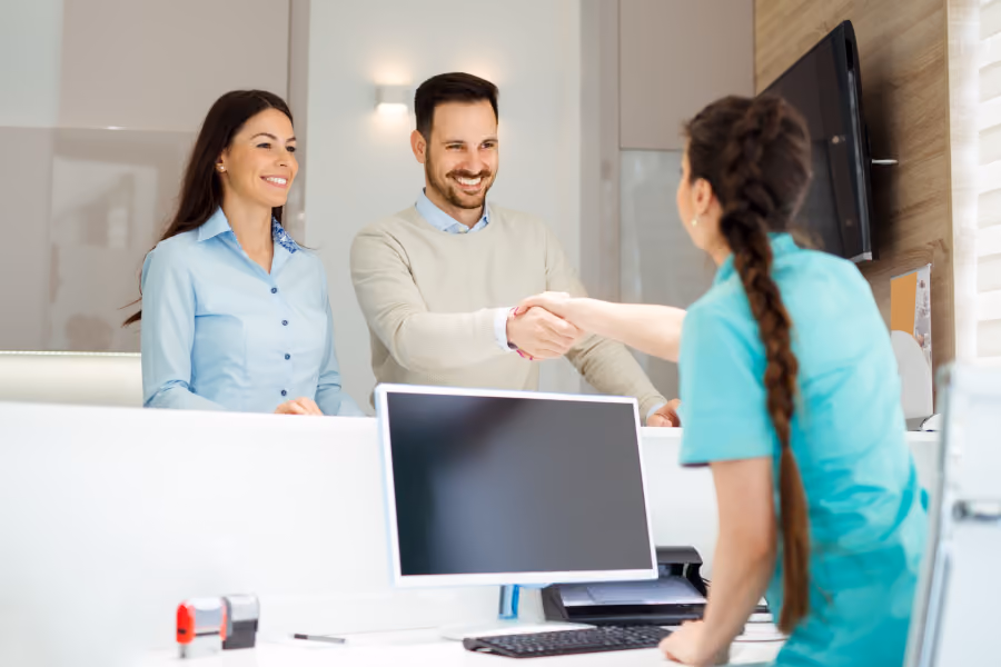 Smiling couple shaking hands with a receptionist in a turquoise uniform at a bright office desk.