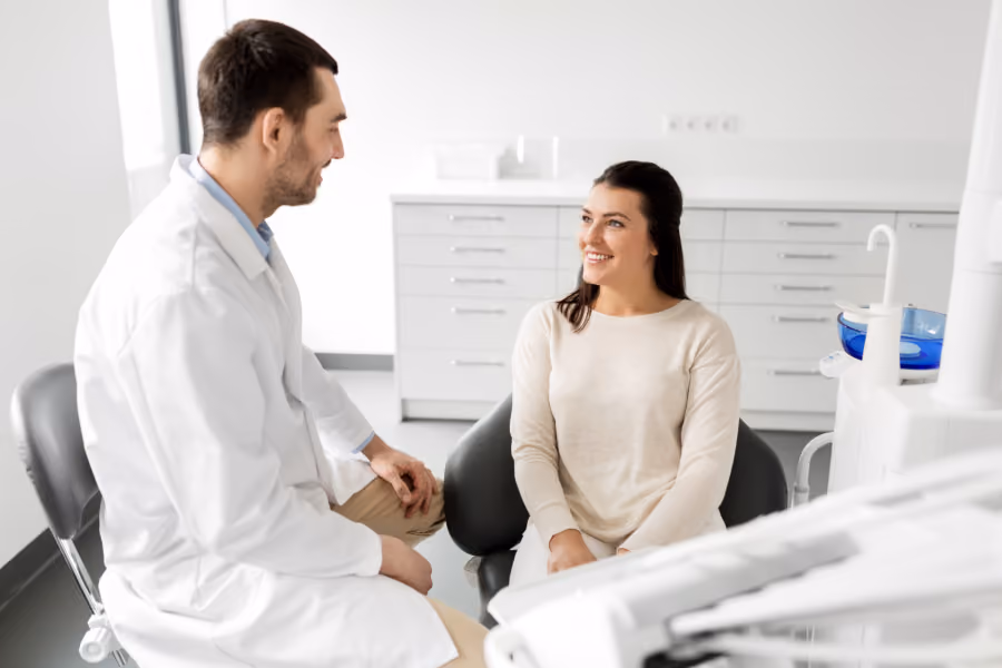 Male doctor in white coat talks with smiling woman seated in a medical office.