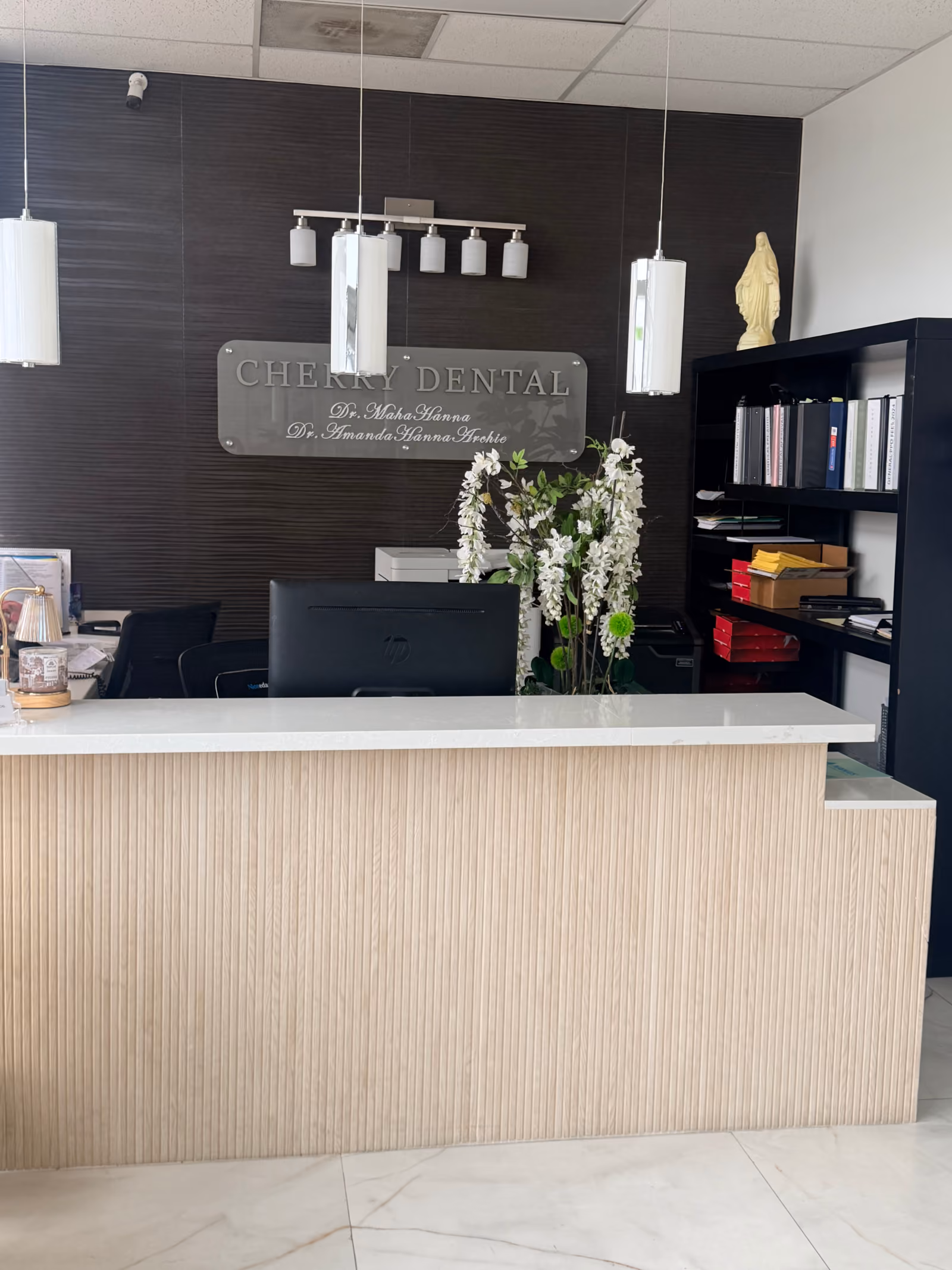 Reception desk area of Cherry Dental with a light wood front panel, white countertop, computer monitor, white flower arrangement, dark wood textured wall behind, and a sign displaying the names Dr. Maha Hanna and Dr. Amanda Hanna Archie.
