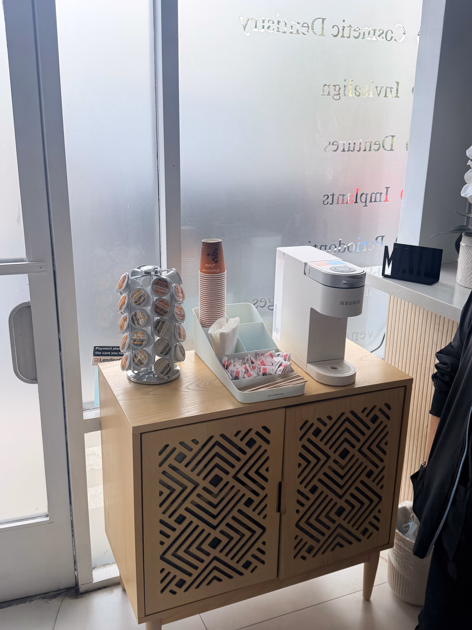 Wooden cabinet with geometric carved doors holding a white Keurig coffee maker, a rotating dispenser of coffee pods, pink disposable cups, and a tray with sugar packets and stirrers near a frosted glass door.