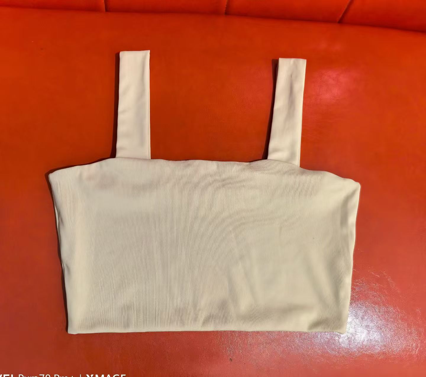 Beige women's ribbed scoop neck crop top with thick shoulder straps laid flat on a red surface.