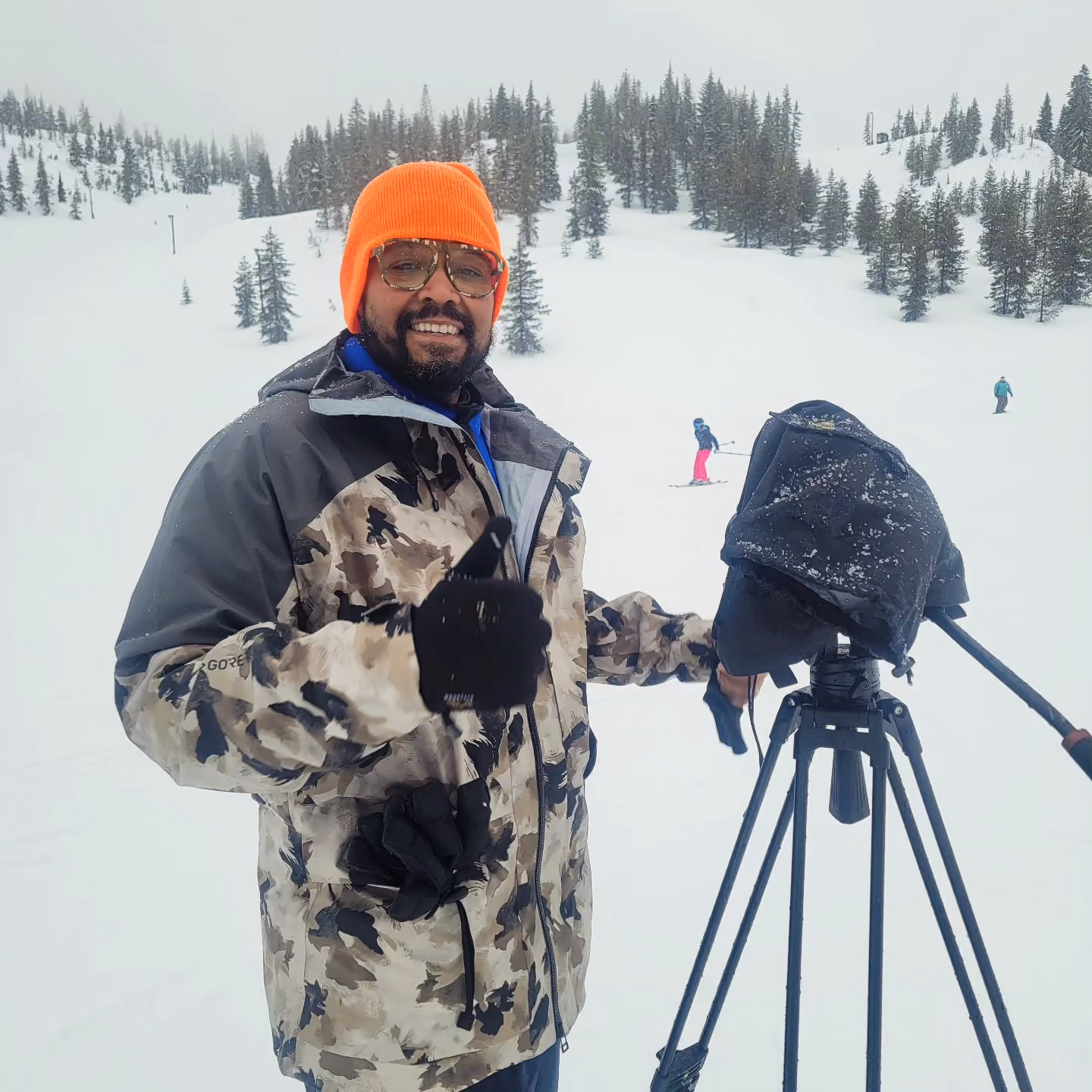 Man in orange beanie and camo jacket with camera tripod in snowy ski area with skiers in background.