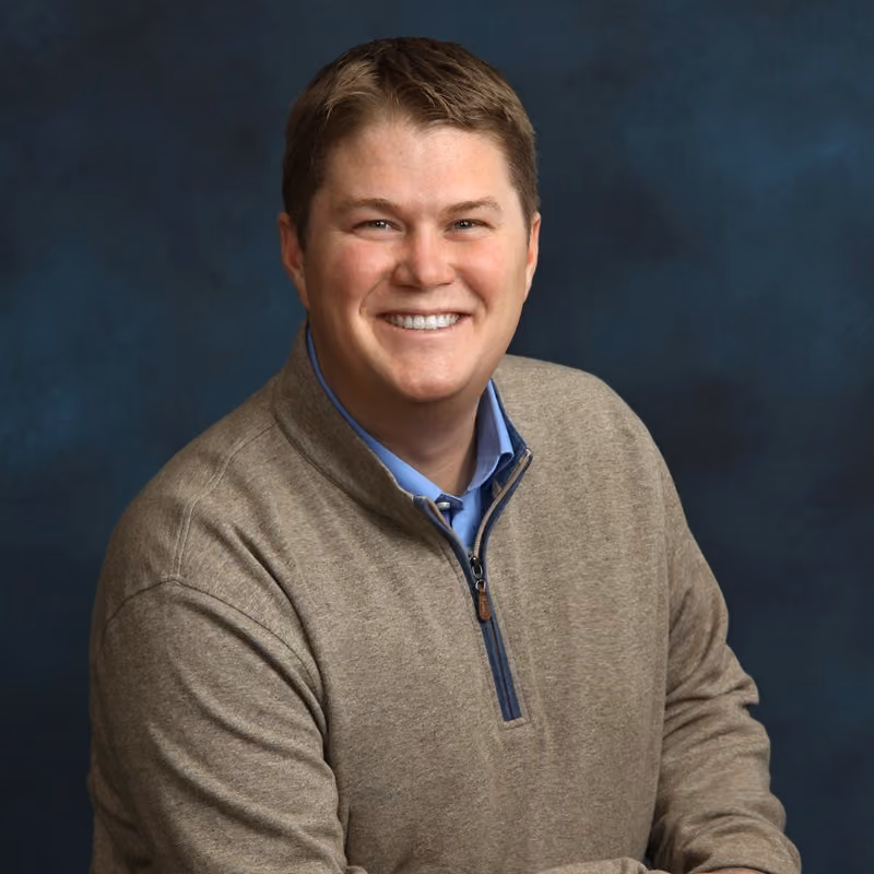 Smiling man in a beige quarter-zip sweater and blue collared shirt against a dark blue background.