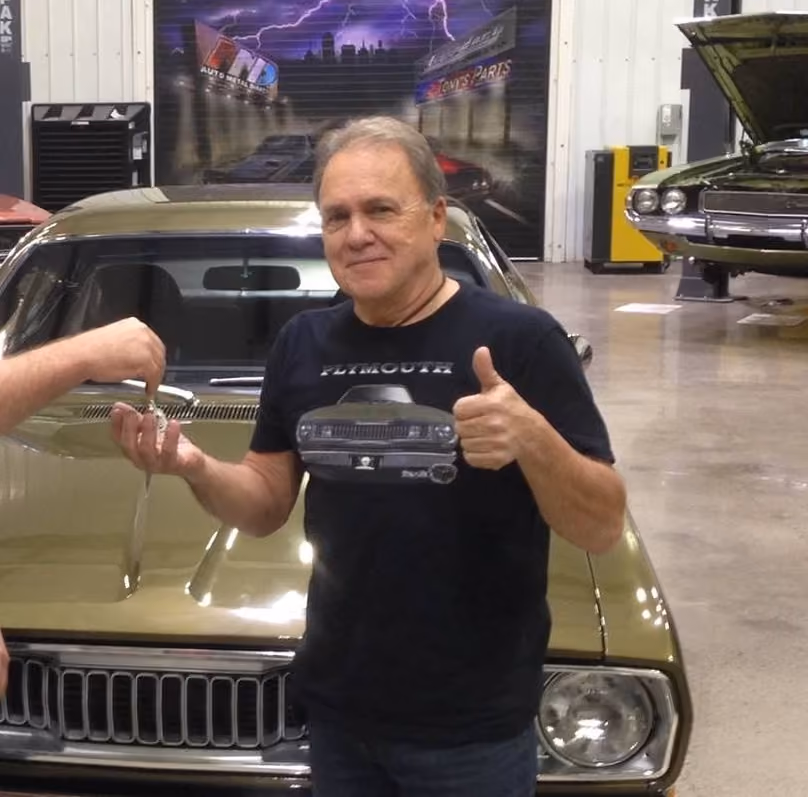 Man wearing a black Plymouth car t-shirt giving thumbs up, standing in front of a classic car indoors.