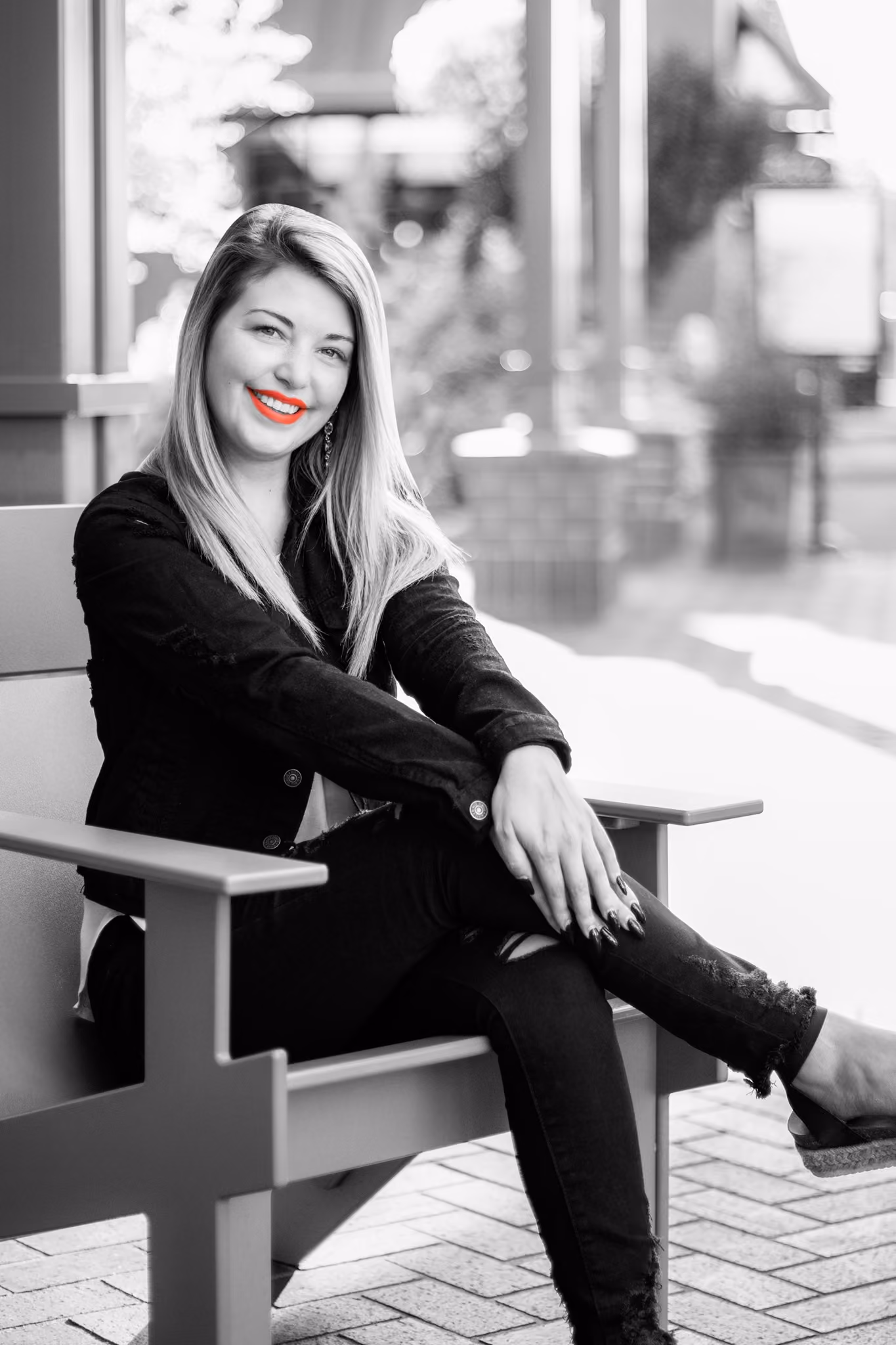 Woman with long hair and red lipstick sitting cross-legged on a bench outdoors.
