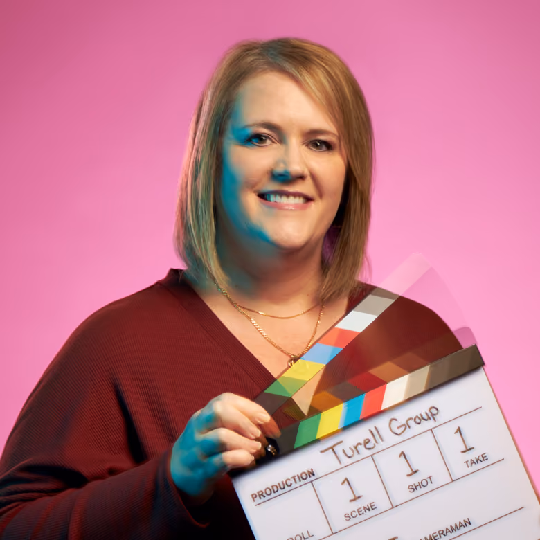Woman smiling wearing maroon top holding a film clapperboard labeled Turell Group on pink background.
