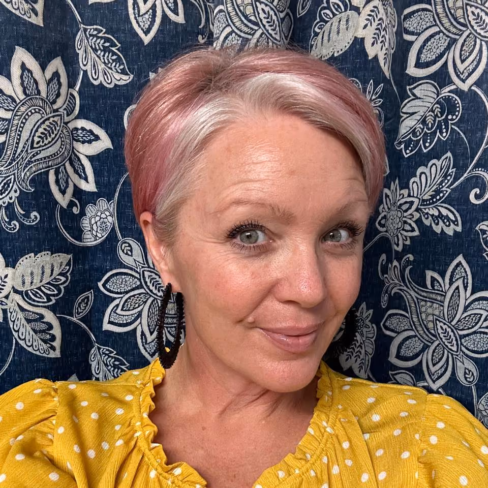 Smiling woman with short pink and white hair, black hoop earrings, yellow polka dot top, floral blue background.