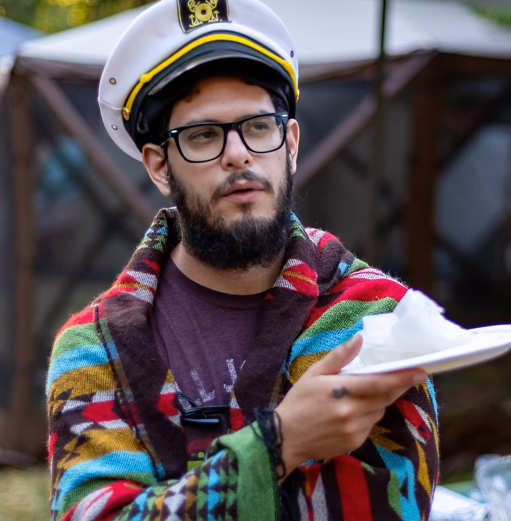 Bearded man in glasses and a captain's hat draped with a colorful blanket holding a white plate.