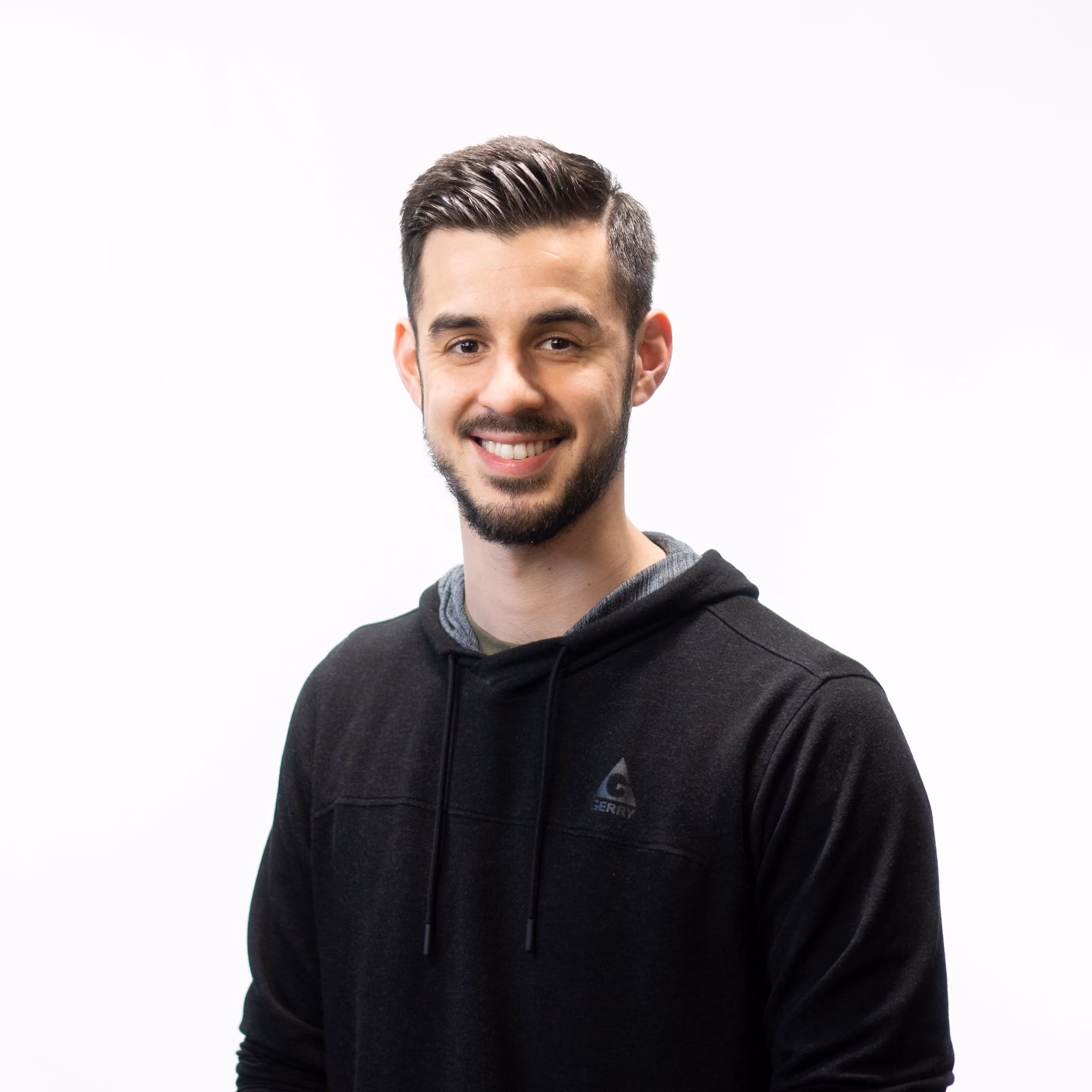 Young man with neatly styled dark hair and beard, smiling, wearing a black hoodie on white background.