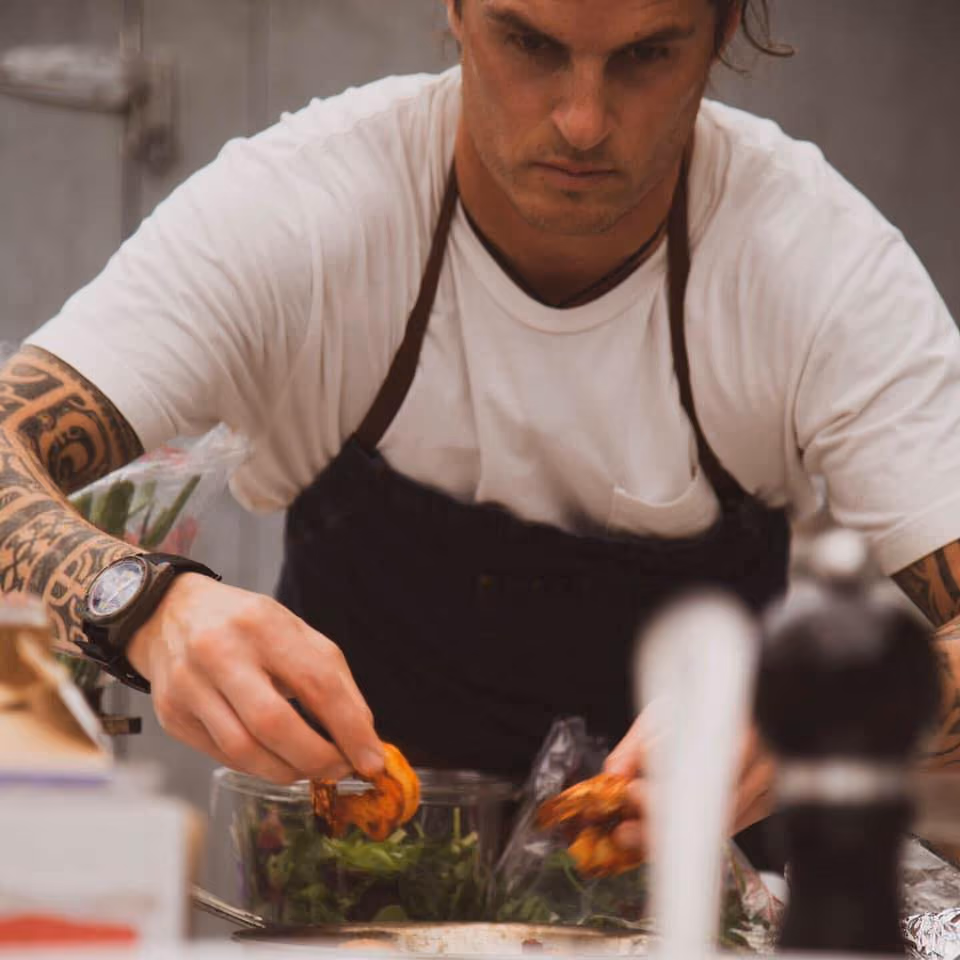 Chef with tattoos and watch placing grilled shrimp on a salad bowl in a kitchen setting.