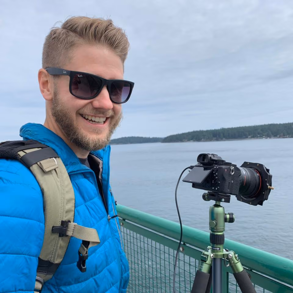 Smiling man wearing sunglasses and blue jacket stands next to camera on tripod by water and green railing.
