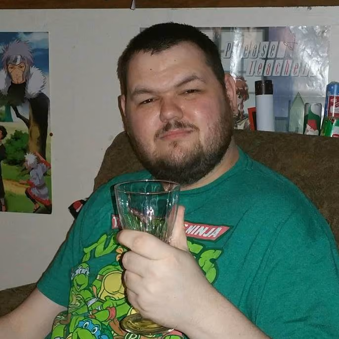 Man with beard in green Teenage Mutant Ninja Turtles shirt holding empty glass, sitting on brown chair.