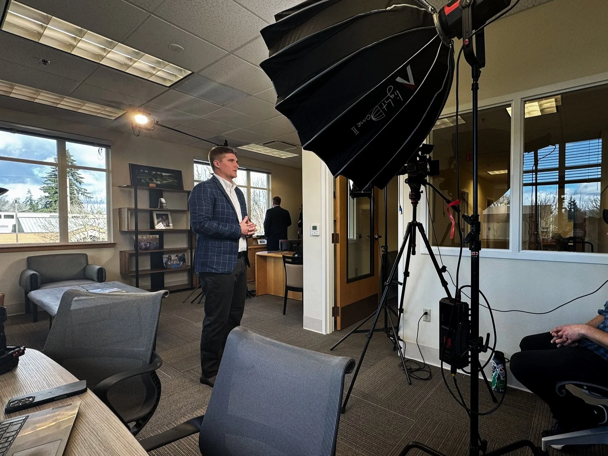 Man in blue checked blazer standing in an office with professional lighting and camera equipment.