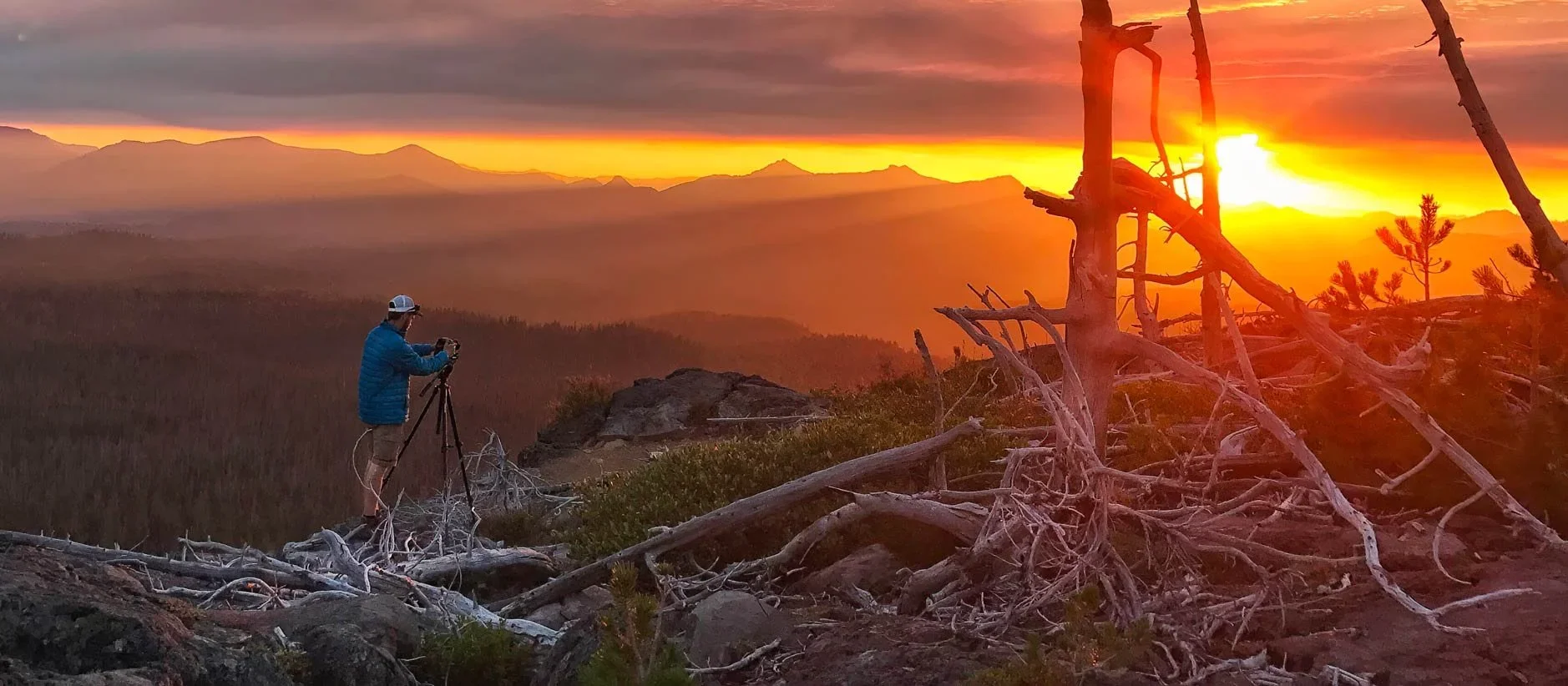 Person adjusting camera on tripod atop a mountain during vibrant orange sunset with tree trunks and rocks nearby.