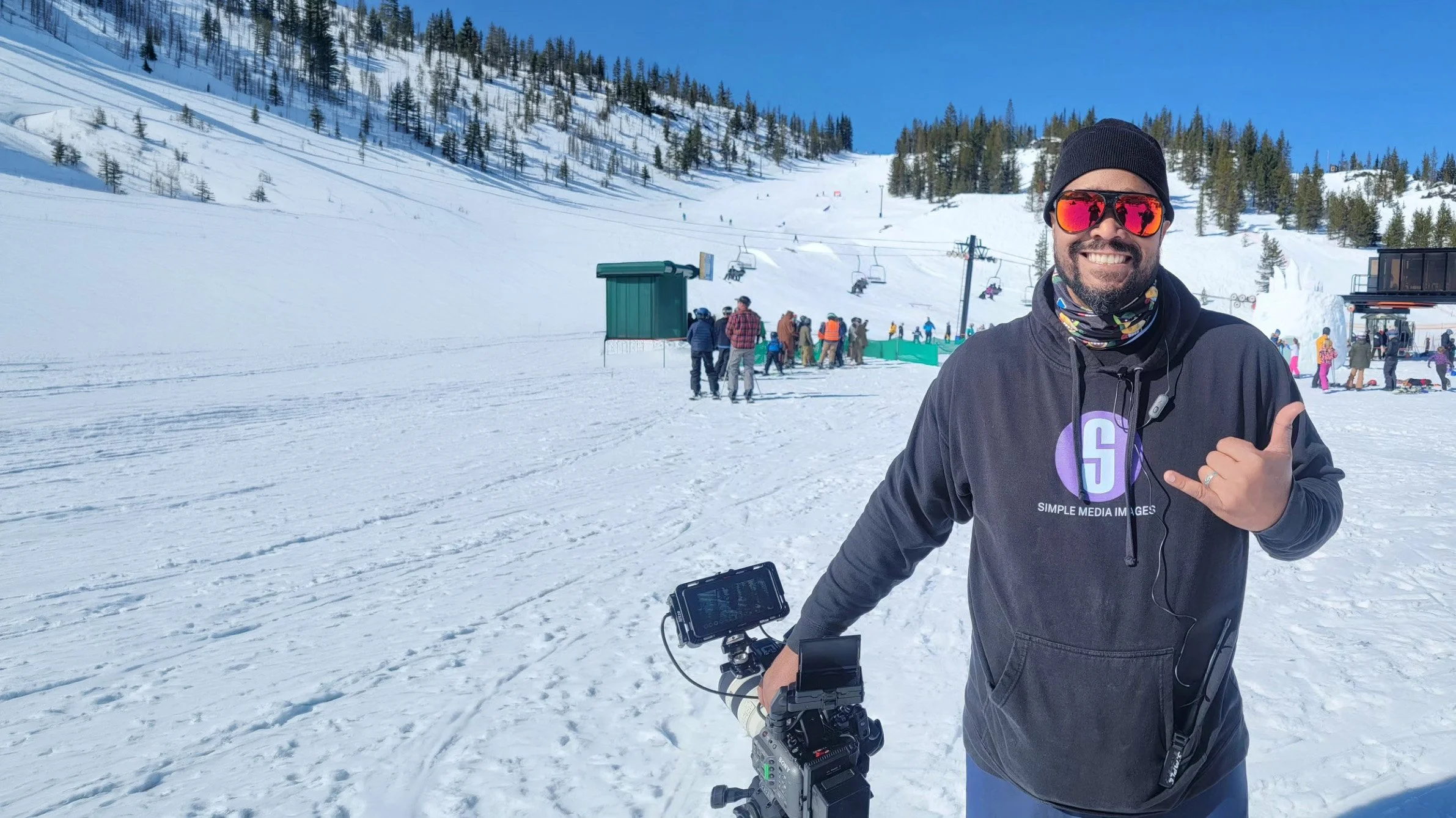 Smiling man in sunglasses and black hoodie stands in snow holding a camera at a ski resort.
