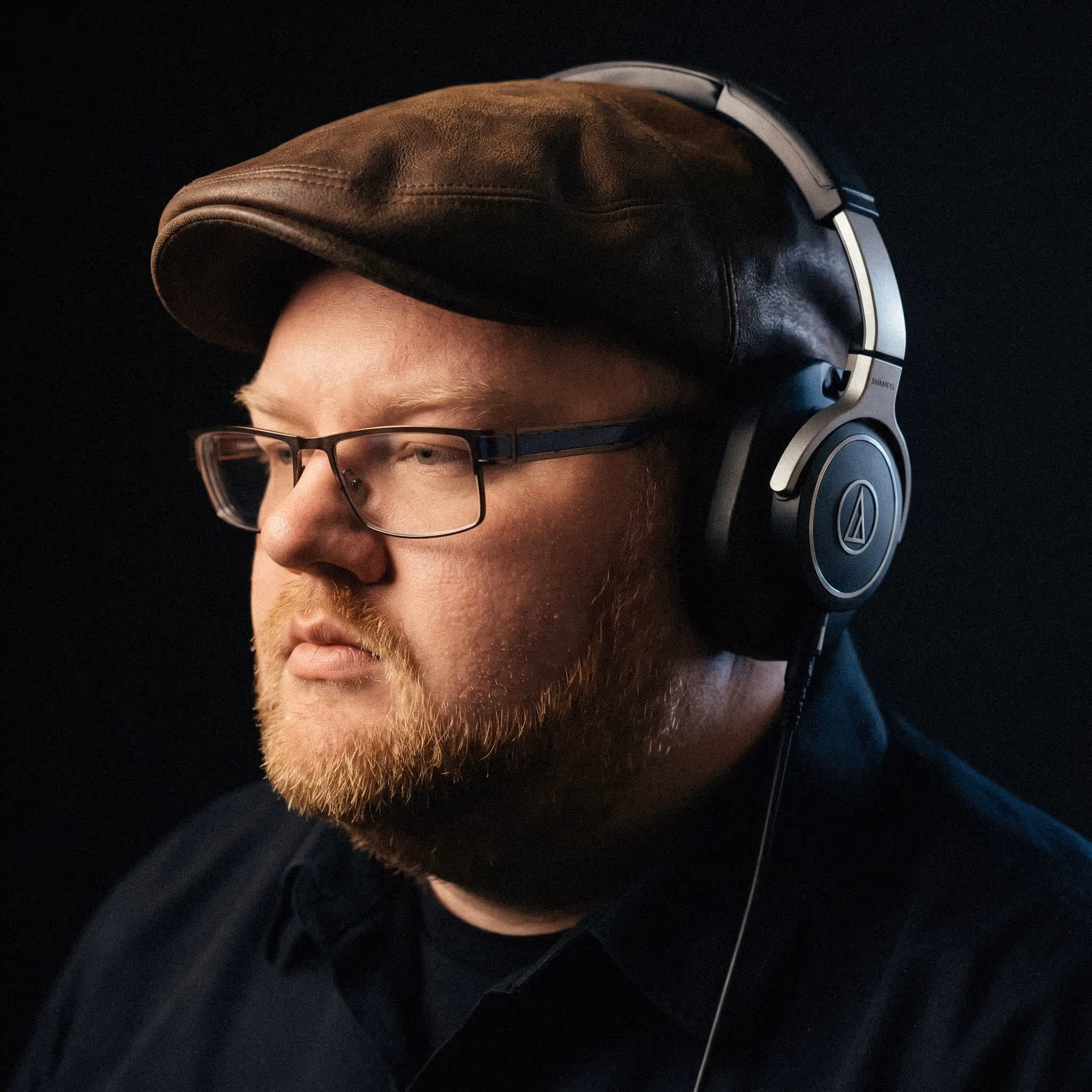 Man with glasses, brown cap, and headphones against a dark background looking to the left.