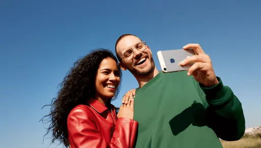 A smiling man and woman outdoors taking a selfie together; the man is holding a smartphone while the woman stands close with her hand on his shoulder against a clear blue sky.