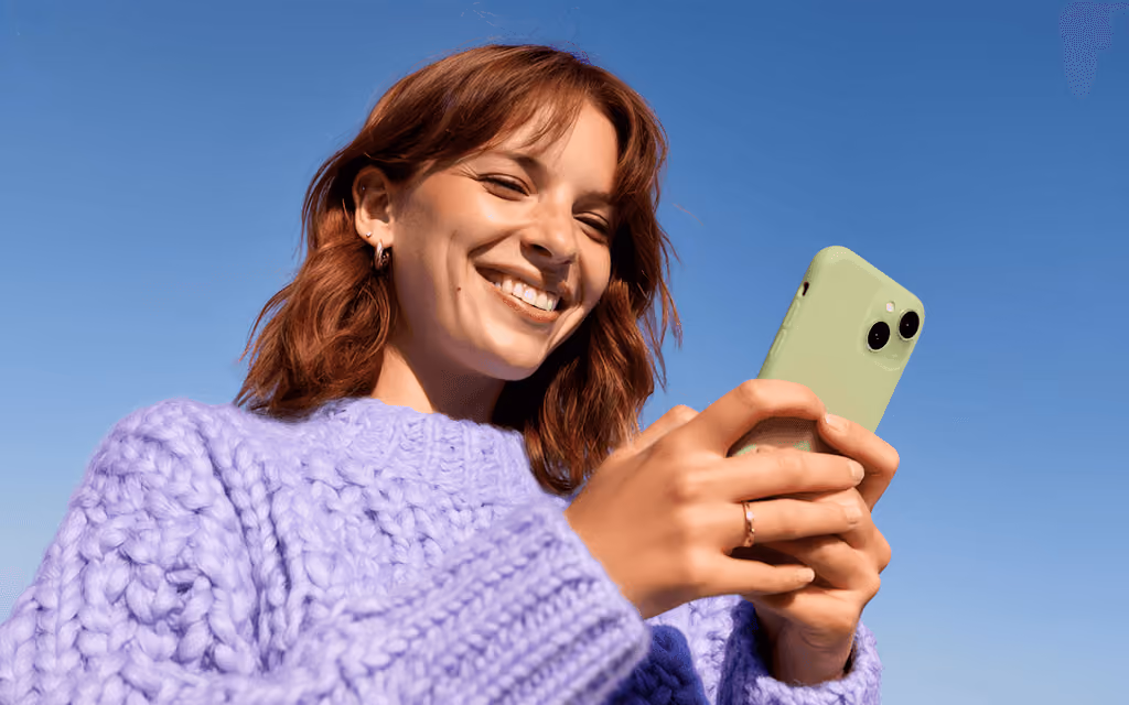 Happy woman with short reddish hair wearing a chunky lavender sweater, smiling and looking at a green smartphone against a clear blue sky.