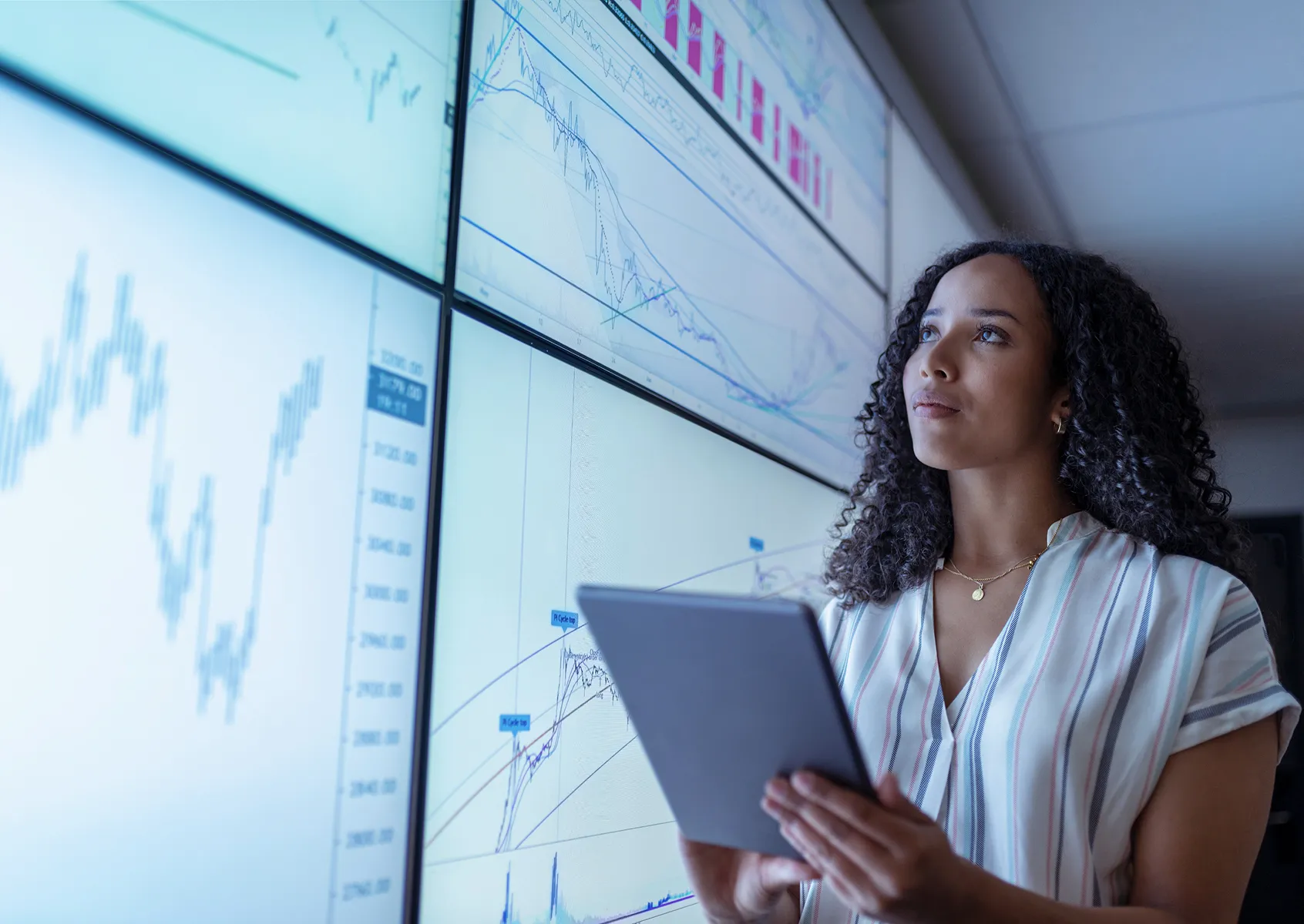 Analyst holding a tablet in front of wall screens showing financial market charts