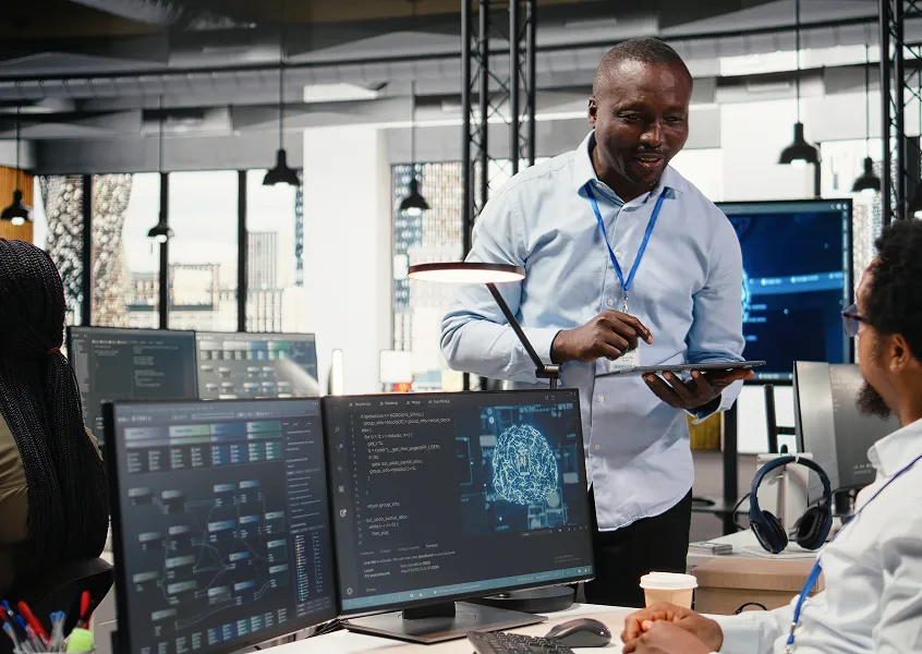 AI engineer holding a tablet beside monitors showing code and an AI brain graphic