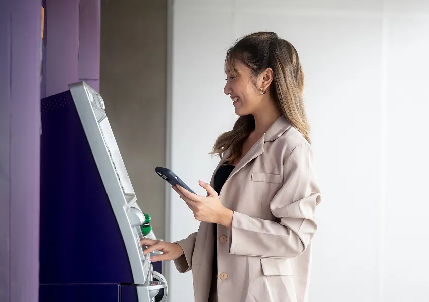 Woman using an ATM while checking her smartphone, representing AI-enabled digital banking in a real-world workflow.