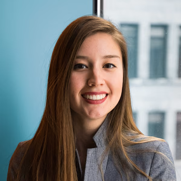 Smiling woman with long brown hair wearing a gray jacket standing by a window with buildings outside.