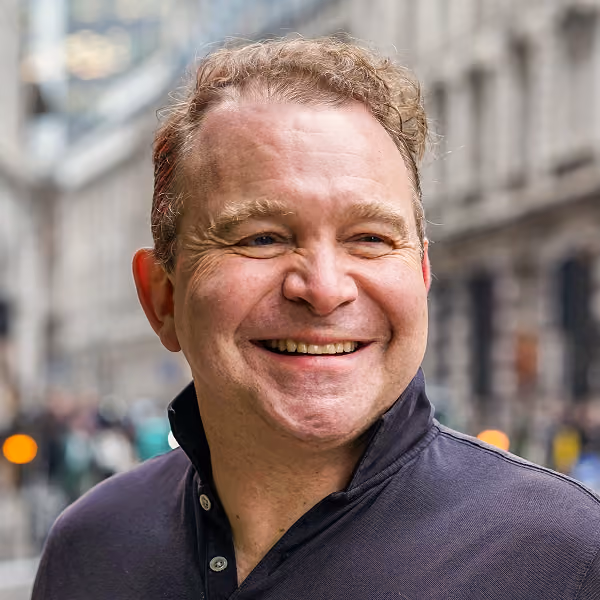 Smiling middle-aged man with curly hair wearing a dark collared shirt in an urban street background.