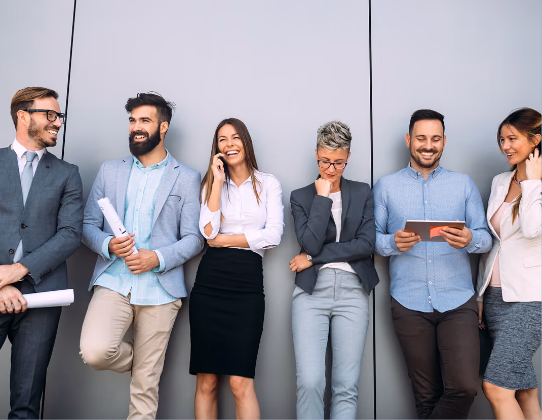 Six diverse professionals standing against a gray wall, smiling and engaging with devices and papers.