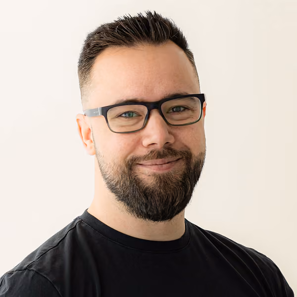 Smiling man with short dark hair, beard, glasses, and a black shirt against a plain light background.