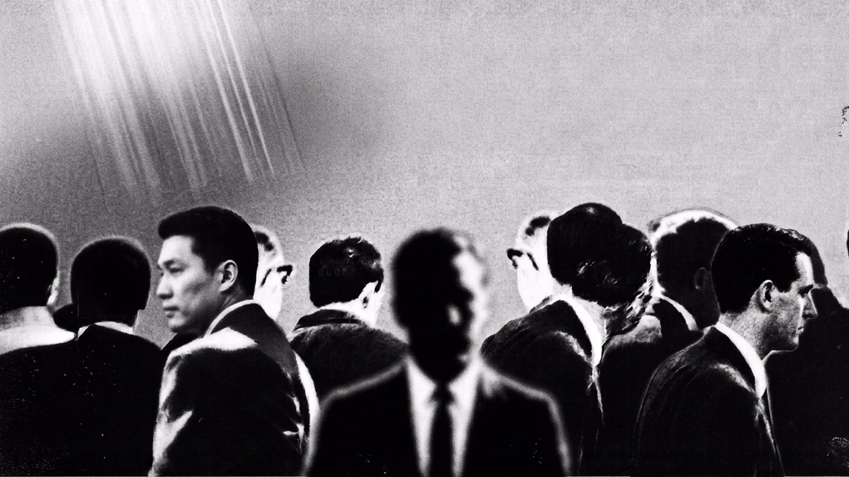Black and white image of men in suits standing or turning in different directions, one blurred in front.